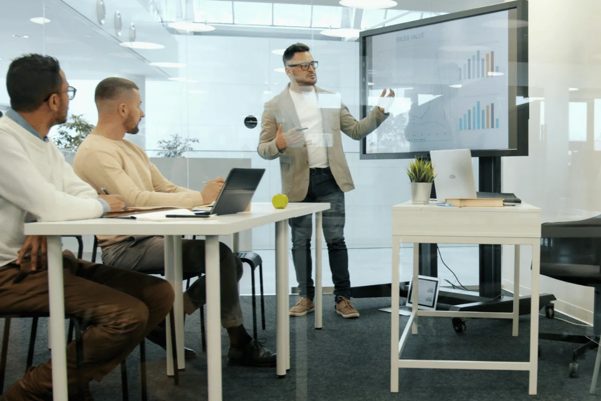 Man in casual blazer presents sales graphs on a screen in a modern office. Three colleagues sit attentively at a table, fostering a focused atmosphere.