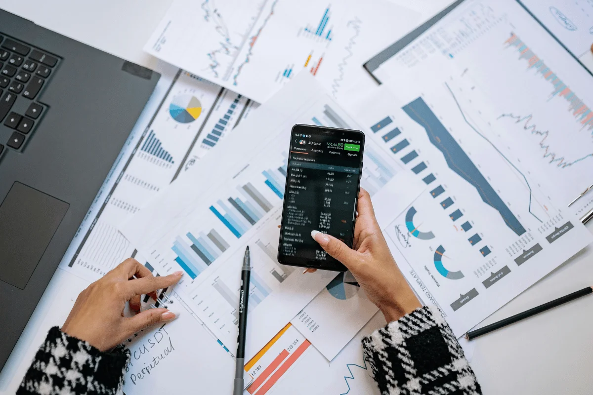Hands analyzing financial charts with a smartphone displaying stock data. Papers and a laptop keyboard are visible, indicating a business setting.
