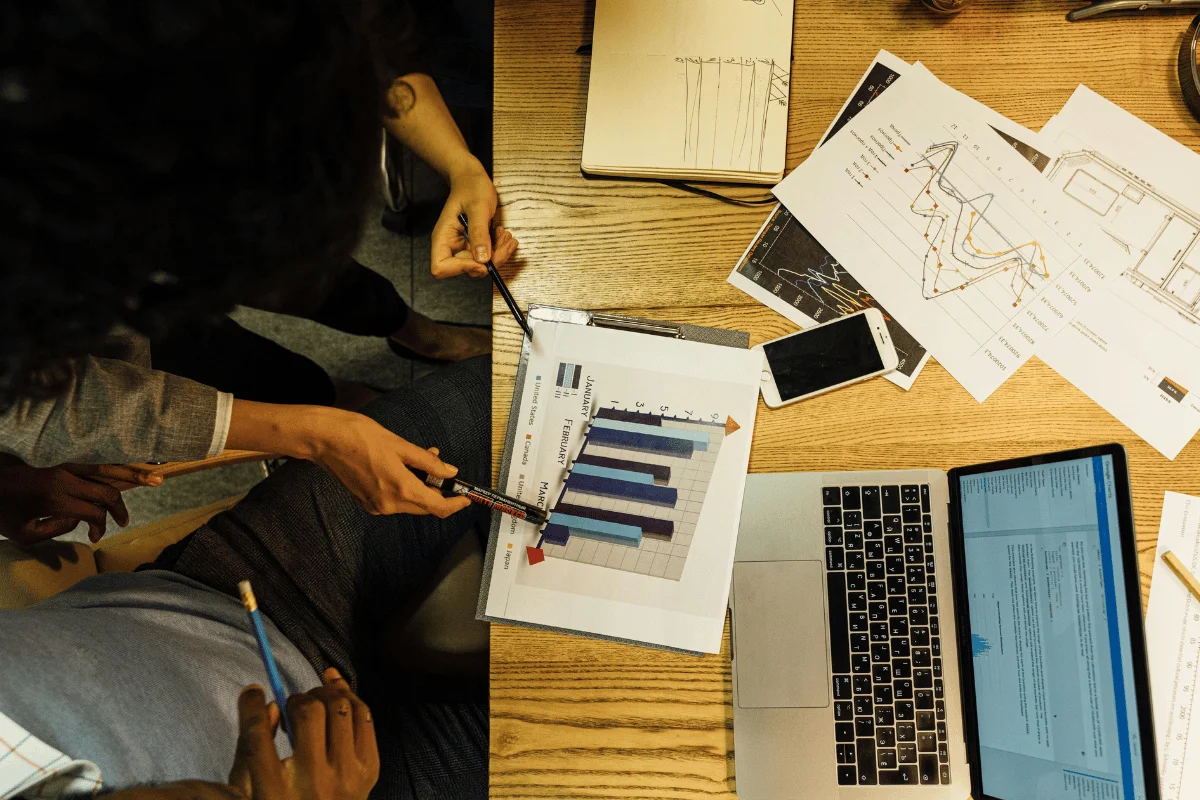 Overhead view of a workspace with people discussing bar charts. A laptop, papers with graphs, and a smartphone are on a wooden table, indicating a business setting.