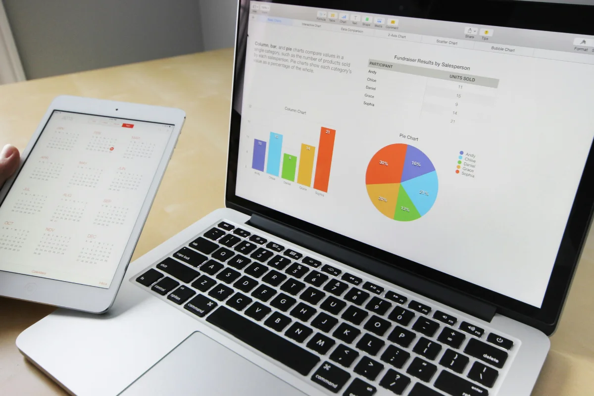 A laptop displaying colorful bar and pie charts sits on a desk. Next to it, a hand holds a tablet showing a calendar, conveying a business-focused setting.